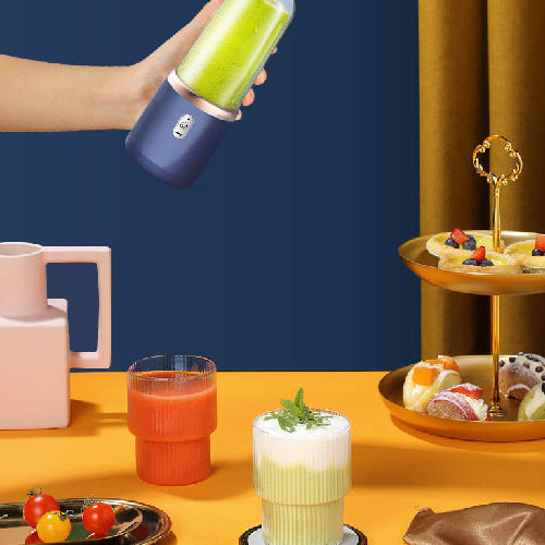 Person holding a blue portable blender with a green smoothie, surrounded by drinks and snacks on a yellow table.
